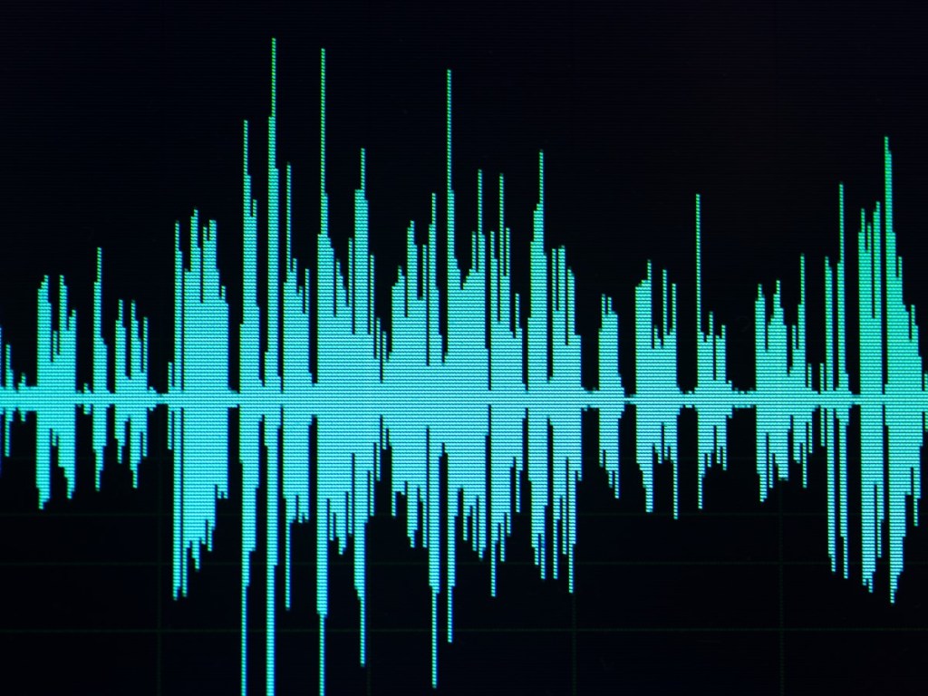 alt=Image shows a visual representation of an audio waveform. The waveform consists of jagged peaks and troughs in turquoise on a black background, indicating varying sound frequencies and aplitudes, reflecting the connection between frequency Hz and well-being